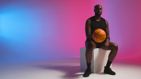 Studio-Portrait-Shot-Of-Seated-Male-Basketball-Bouncing-Ball-Against-Pink-And-Blue-Lit-Background